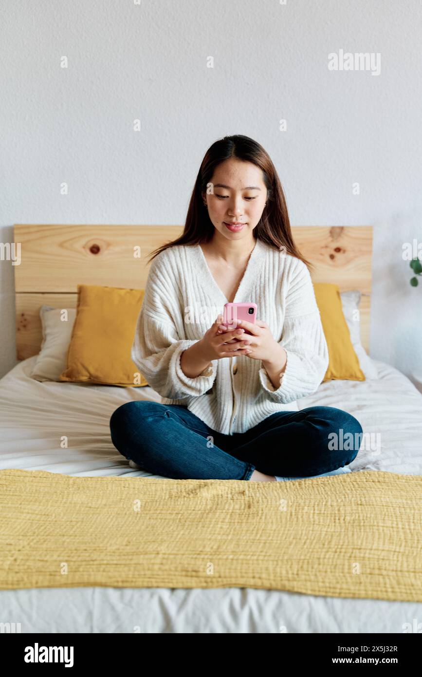 Una donna seduta su un letto e che usa il suo smartphone Foto Stock