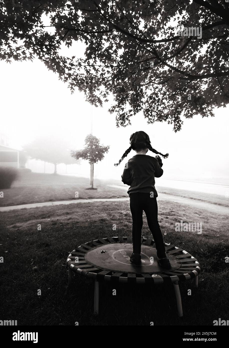 Giovane ragazza con trecce che salta sul trampolino sotto l'albero Foto Stock