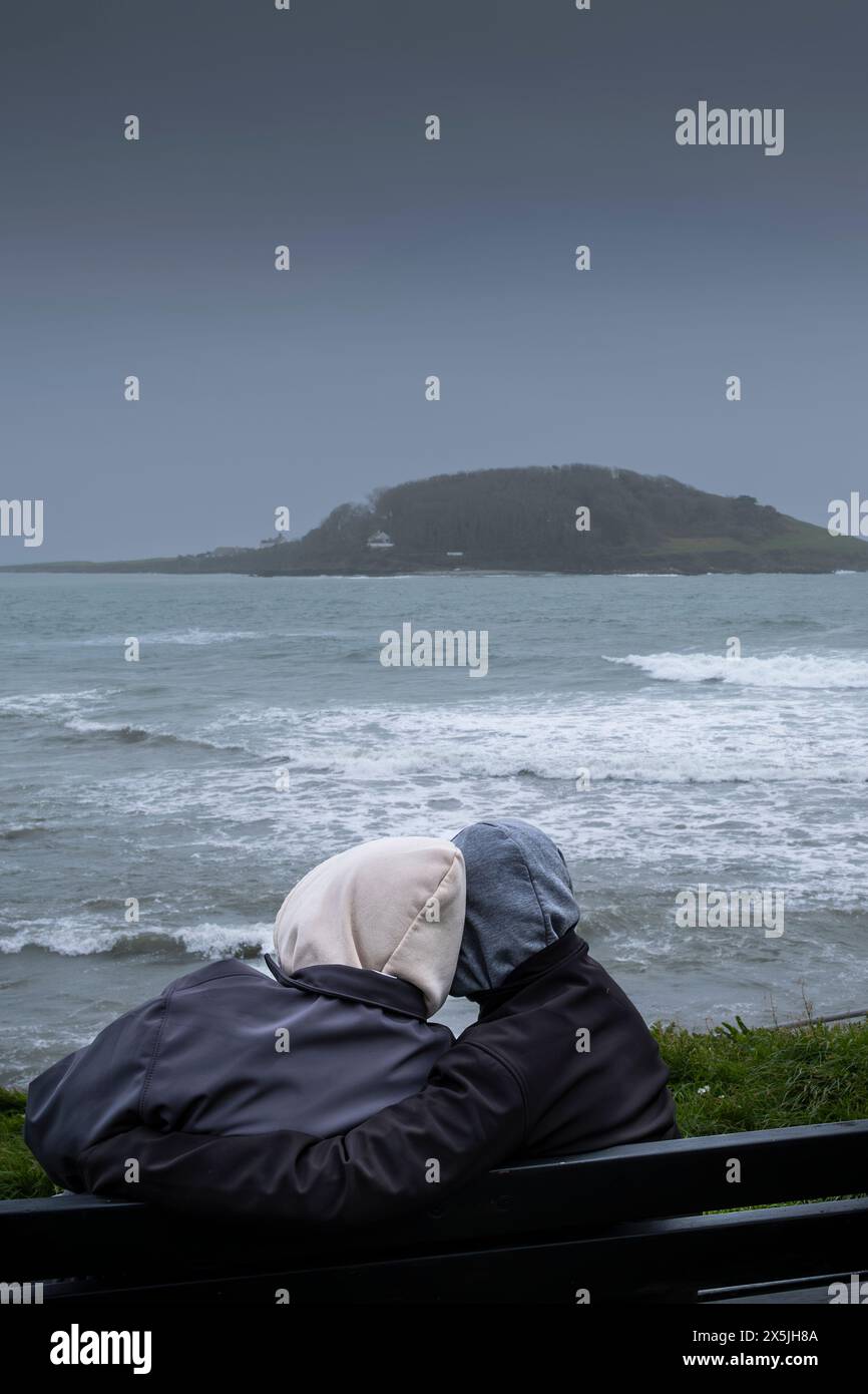 Meteo per il Regno Unito. Due persone che abbracciano e si siedono su una panchina che si affaccia sul mare verso la storica Looe Island al largo della costa della Cornovaglia nel Regno Unito. Foto Stock