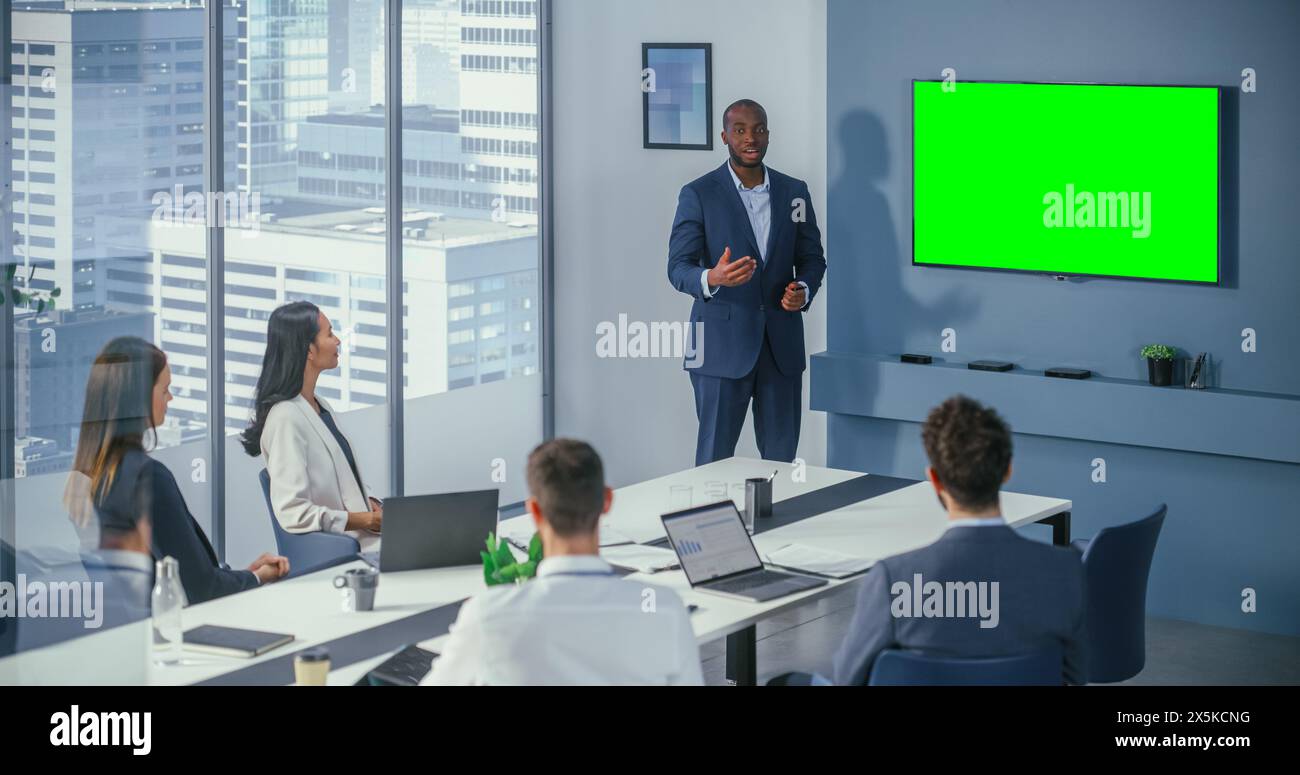 Riunione di diverse sale riunioni per uffici: Il Project Manager Black male utilizza la TV a parete Chroma Key Green Screen per presentare il prodotto al gruppo di investitori. Strategia di e-commerce. Shot statico ampio Foto Stock