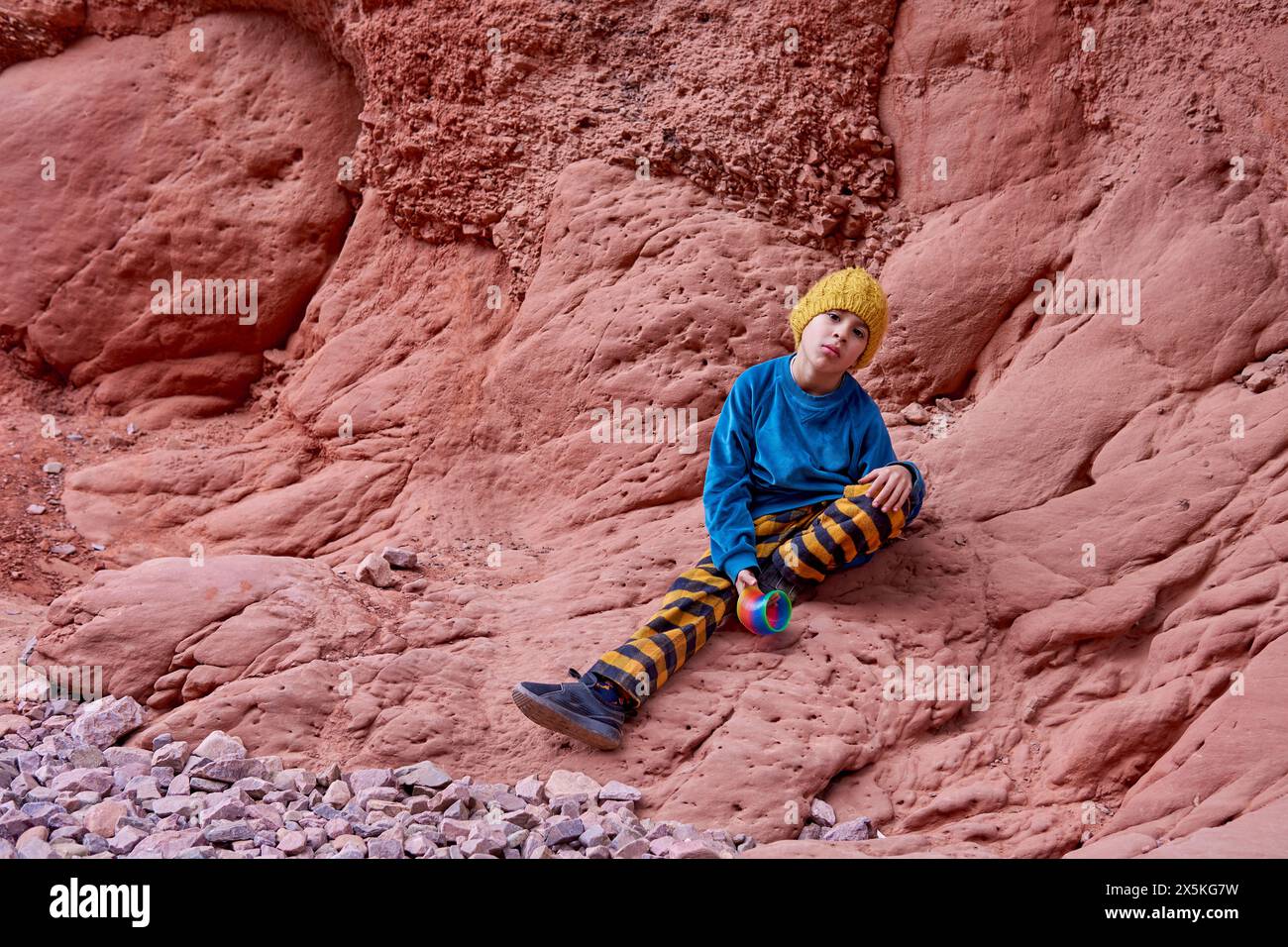 Ragazzo latino seduto su una formazione rocciosa che sembra annoiato dalla macchina fotografica durante un tour di vacanza invernale a la Quebrada de las Señoritas a Jujuy, Argentina. Foto Stock