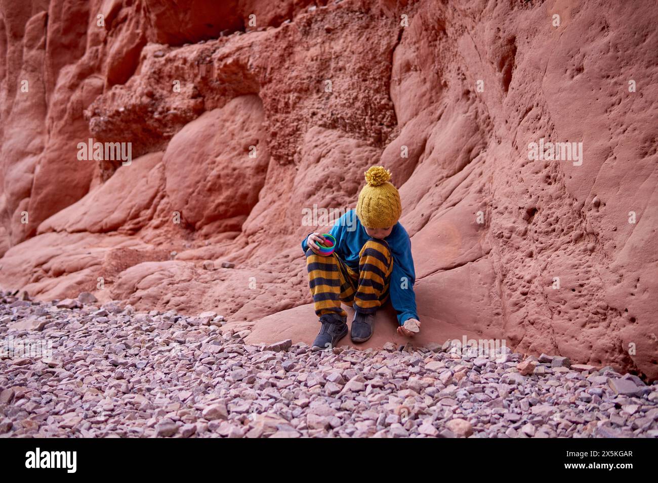 Ragazzo latino seduto su una montagna rocciosa che guarda giù raccogliendo ciottoli durante le vacanze invernali a la Quebrada de las Señoritas a Jujuy, Argentina. Foto Stock