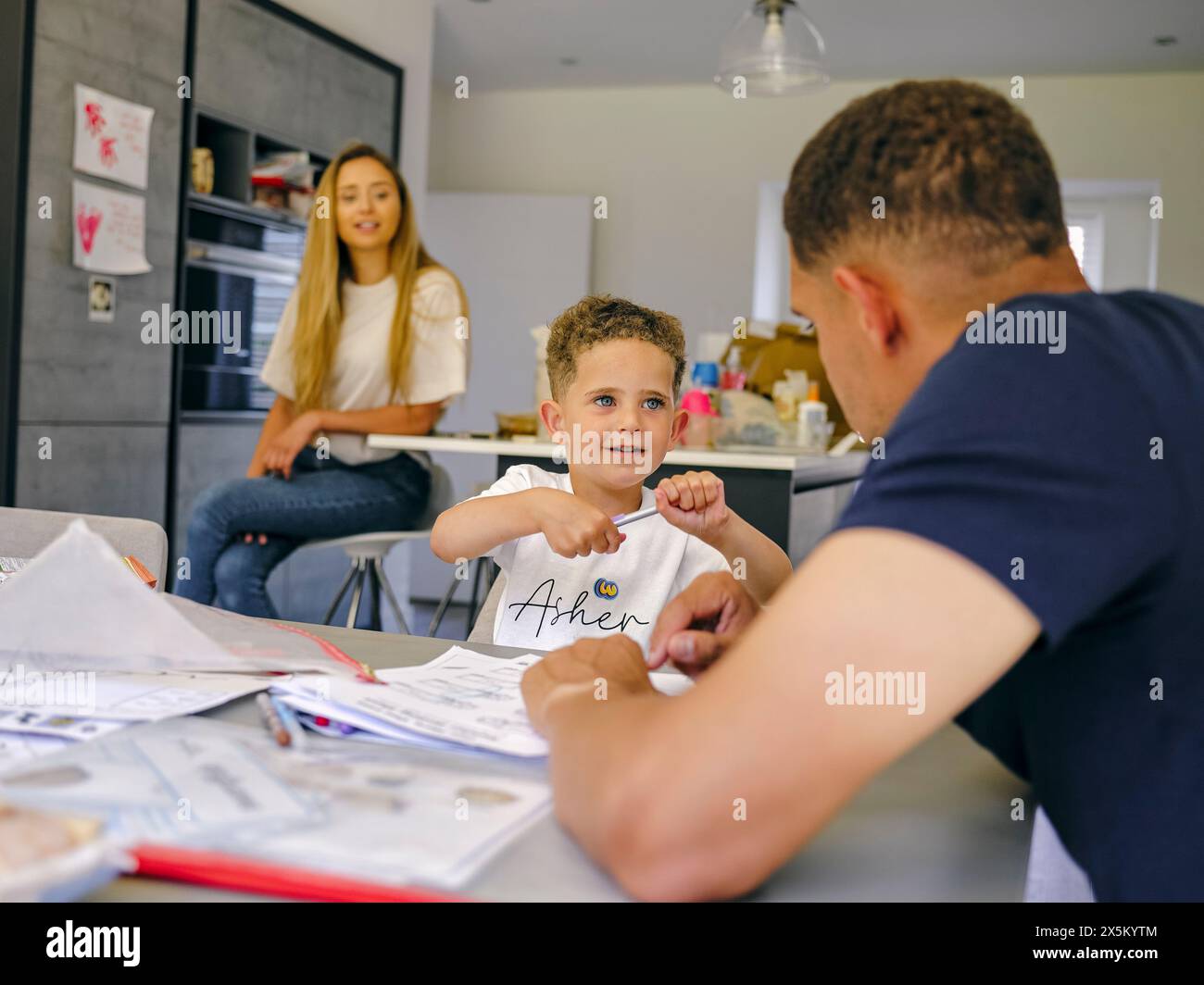 Padre che assiste il figlio a fare i compiti a casa, madre in background Foto Stock