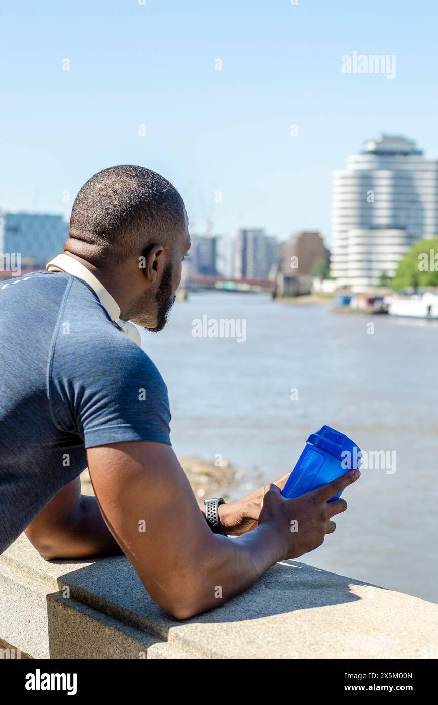 Uomo che tiene in mano un contenitore d'acqua di plastica e guarda il fiume della città Foto Stock