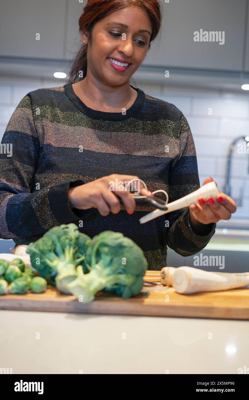 Donna sorridente che sbuccia la radice di prezzemolo in cucina Foto Stock