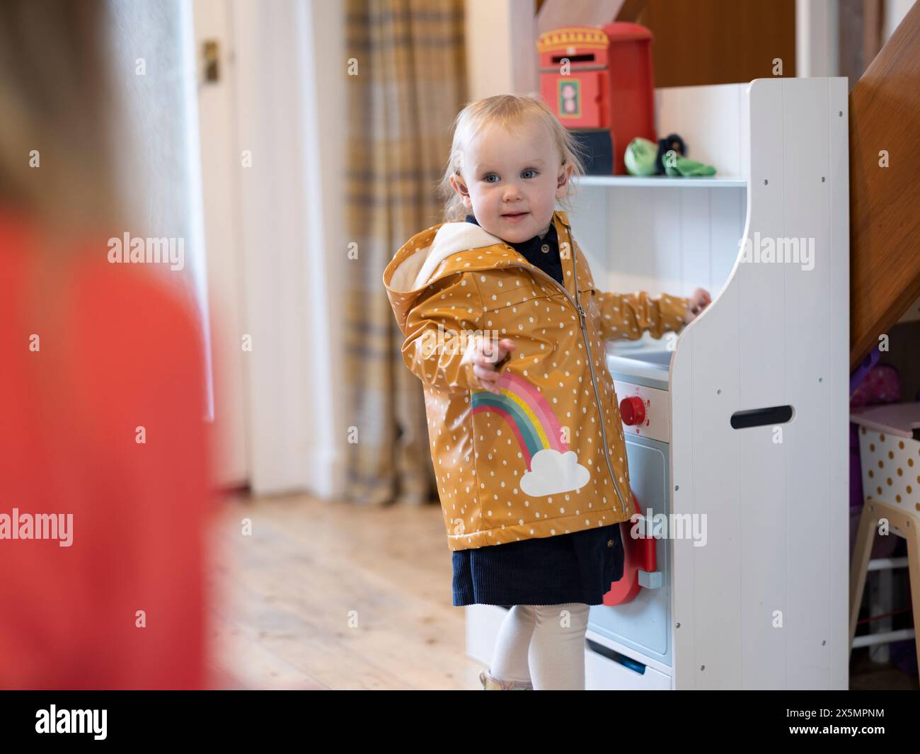 Ragazza bionda che indossa apparecchi acustici e impermeabile accanto alla cucina giocattolo a casa Foto Stock