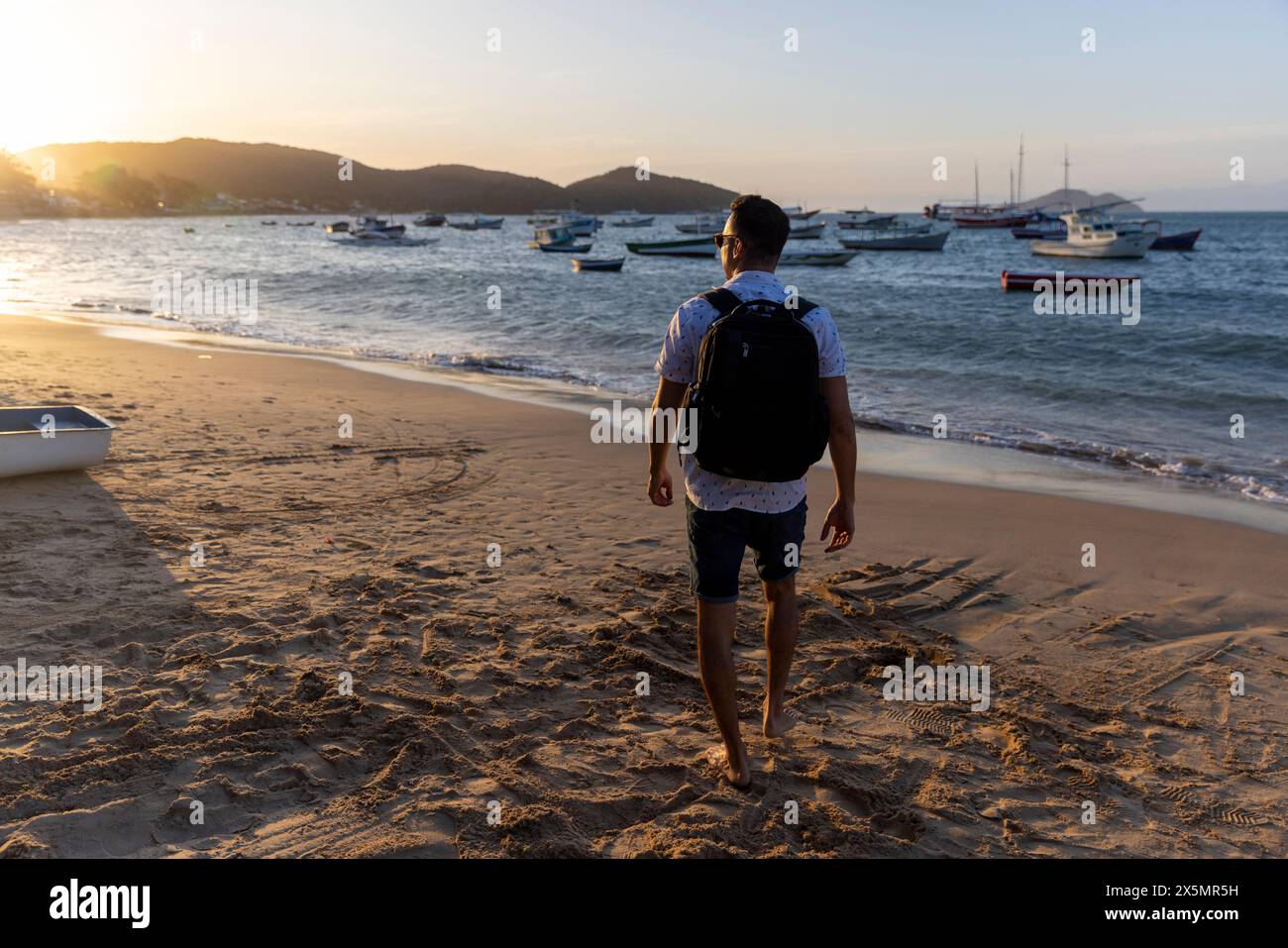Vista posteriore dell'uomo con zaino sulla spiaggia al tramonto Foto Stock