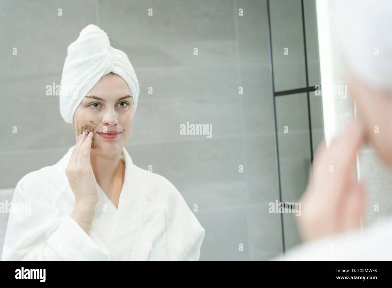 Donna in asciugamano turbante che applica crema esfoliante sul viso in bagno Foto Stock