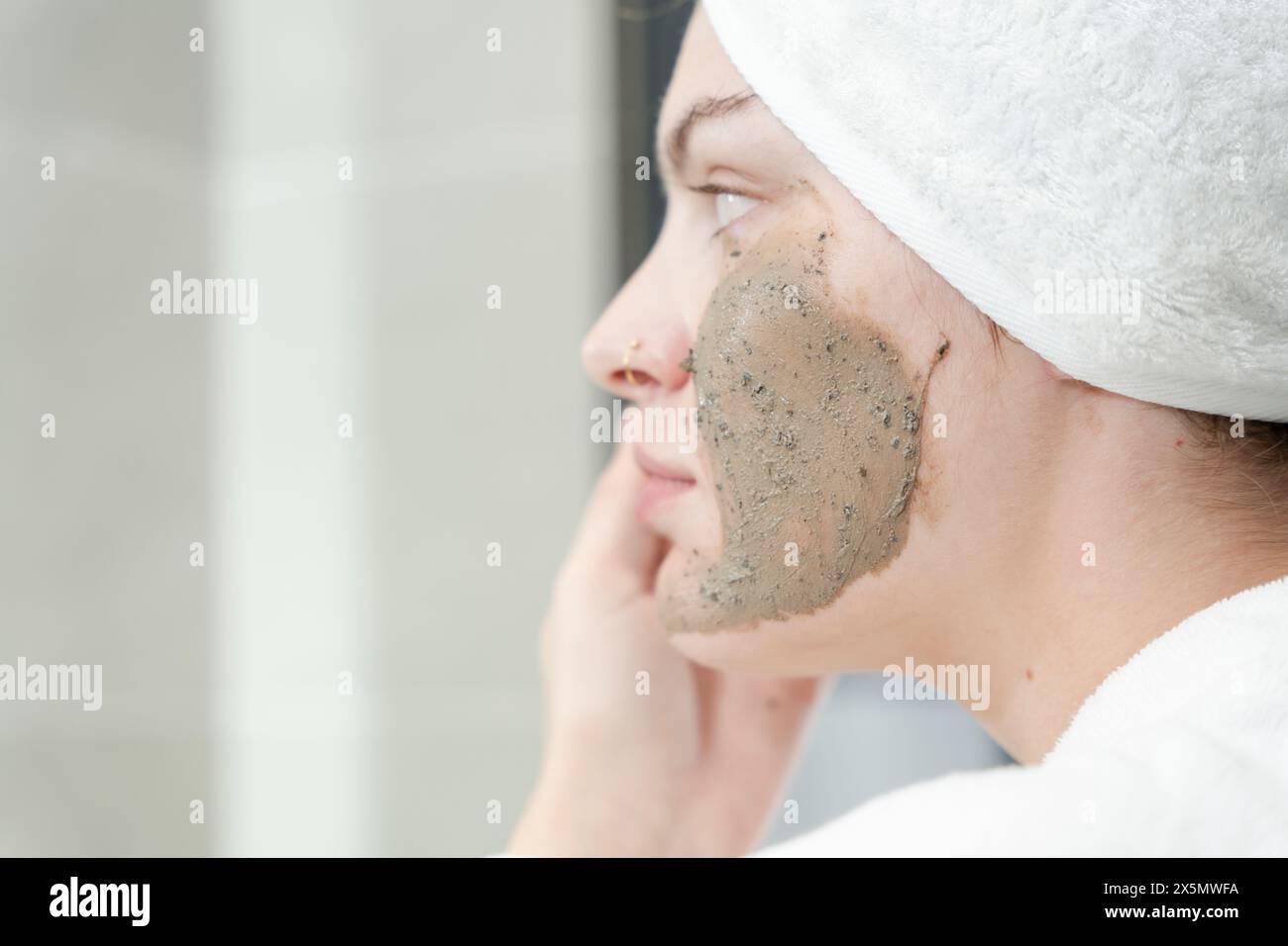 Donna in asciugamano turbante che applica crema esfoliante sul viso in bagno Foto Stock