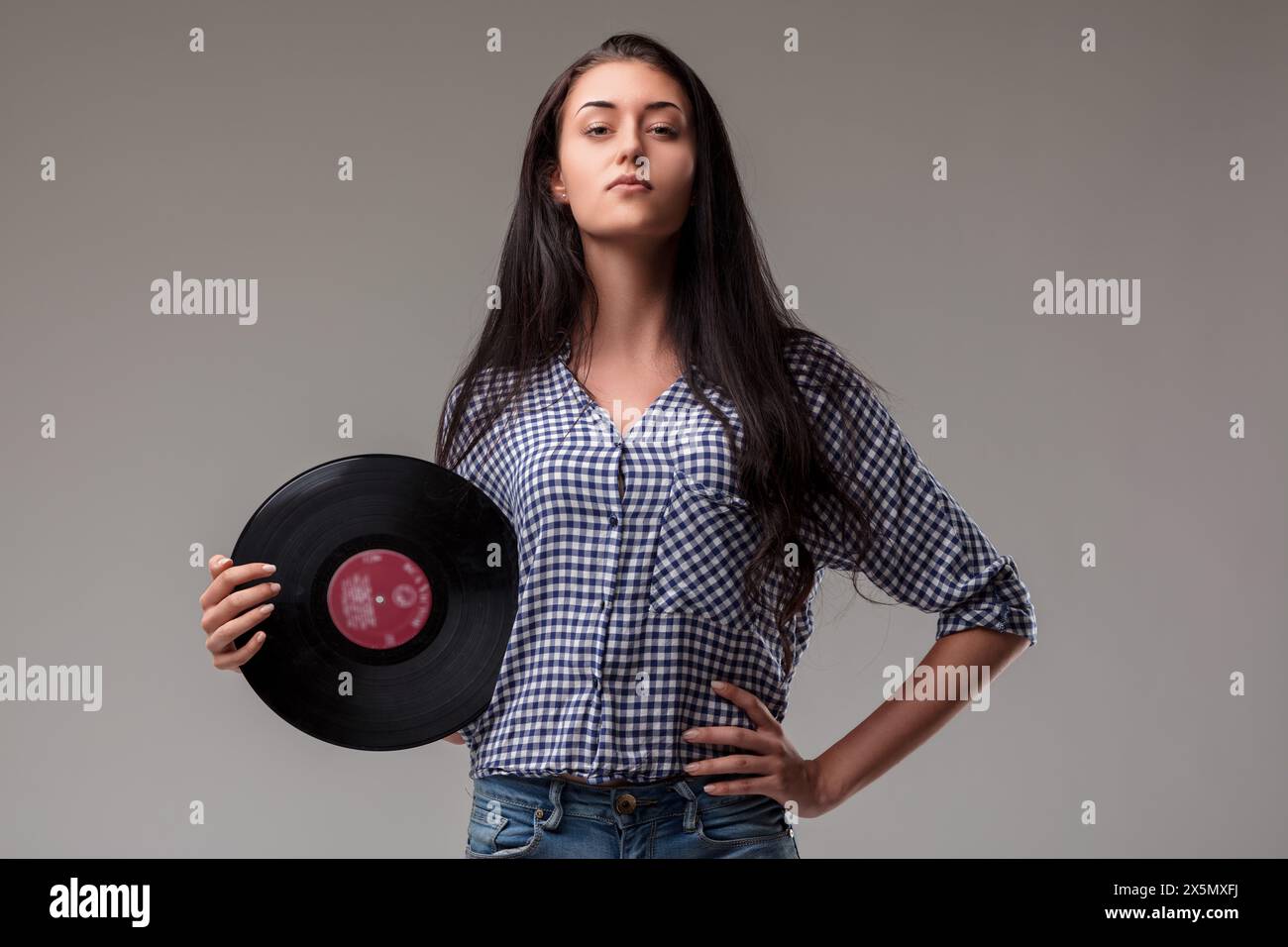 Una giovane donna sicura di sé con una camicia blu Vichy ha un record in vinile, trasudando un comportamento serio Foto Stock