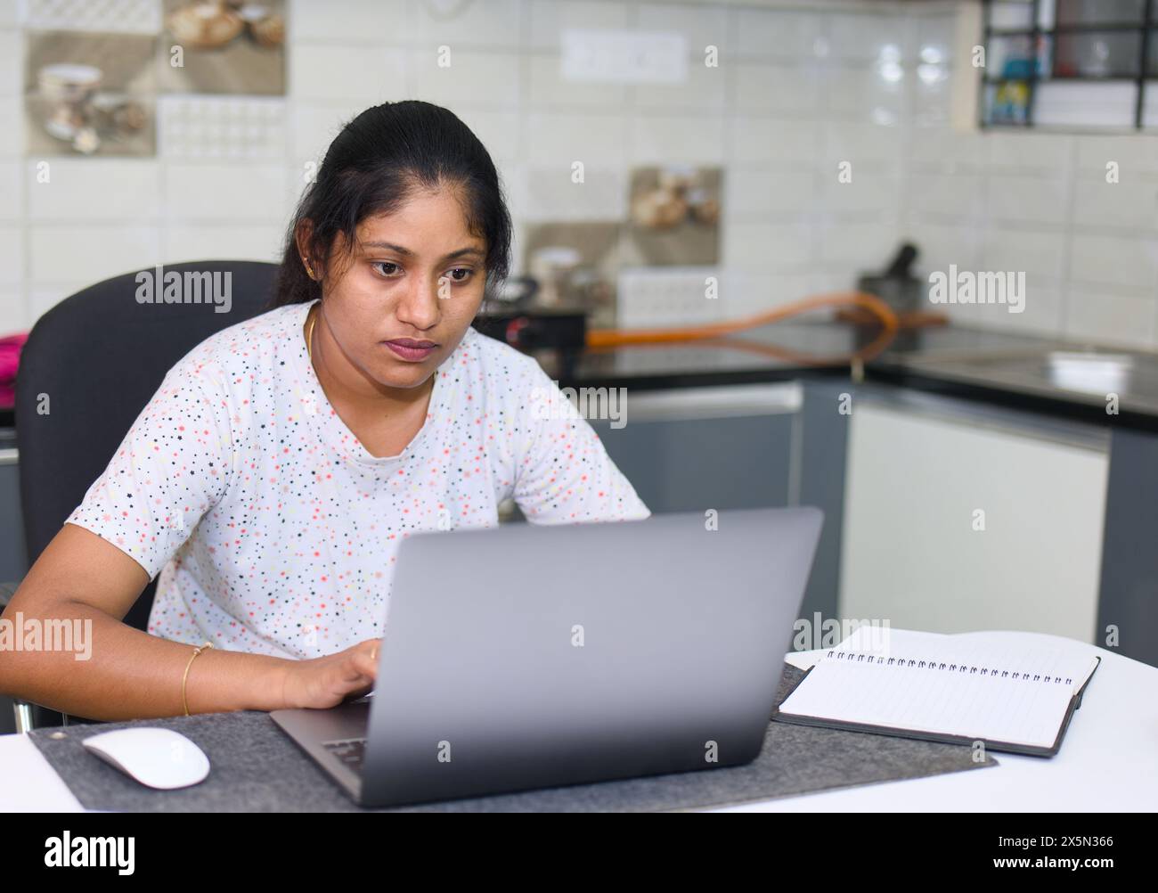 Una donna indiana concentrata affronta in modo efficiente il suo carico di lavoro su un computer portatile mentre fa riferimento a appunti in un notebook al banco della sua cucina. Foto Stock