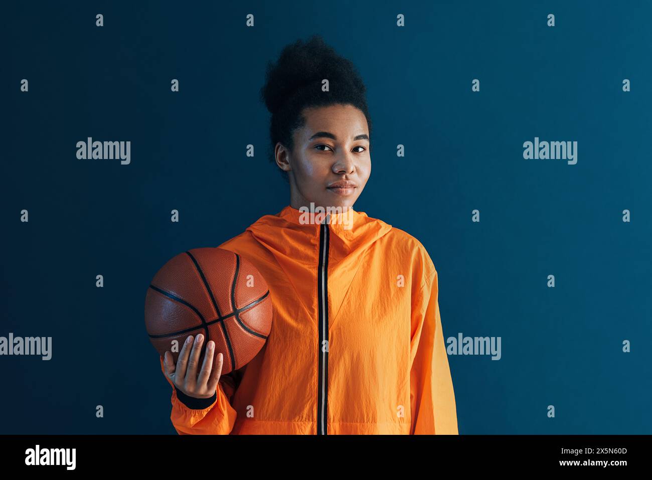 Ritratto di una giovane giocatrice di basket sullo sfondo blu. Donna in abbigliamento sportivo arancione che tiene in mano una pallacanestro che guarda la macchina fotografica. Foto Stock