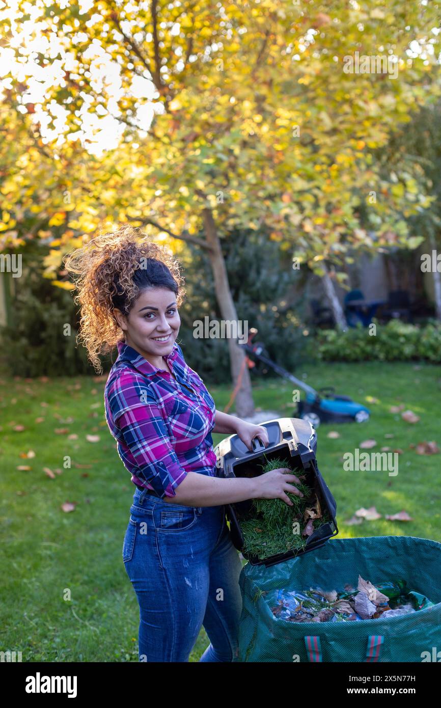 Bella giovane donna che getta erba e foglie dal cestino del rasaerba in giardino Foto Stock