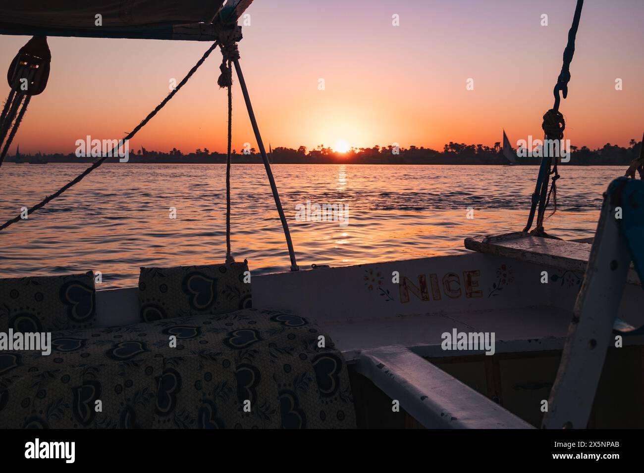 Tramonto sul Nilo visto da una barca con cuscini a fantasia, calme acque ondulate e un cielo arancione brillante che si riflette sulla superficie del fiume. Foto Stock