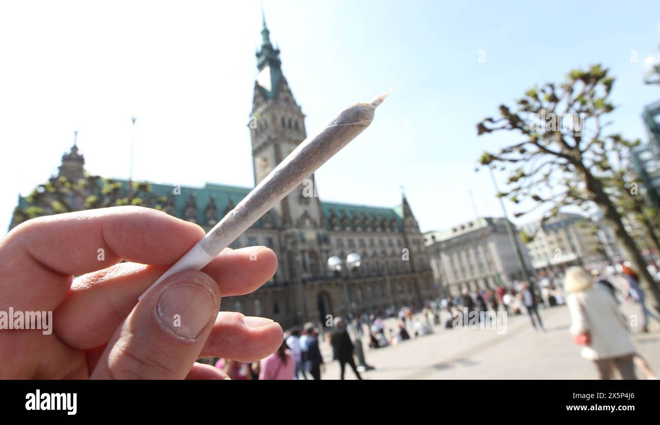 Ein Mann hält einen Joint in der Hand. Im Hintergrund das Hamburger Rathaus. Der hamburger Senat Hat einen Katalog von Bußgeldern für Gesetzesverstöße erlassen, der hohe Geldstrafen und sogar Haftstrafen für Personen vorsieht, die mehr als die erlaubten 25 Gramm Haschisch oder Marihuana auf der Straße dabeihat oder 50 Gramm in der Wohnung besitzt. Altstadt Amburgo *** Un uomo tiene in mano un comune il Municipio di Amburgo sullo sfondo il Senato di Amburgo ha emesso un catalogo di multe per violazioni della legge, che prevede multe elevate e persino pene detentive per le persone che portano più di Foto Stock