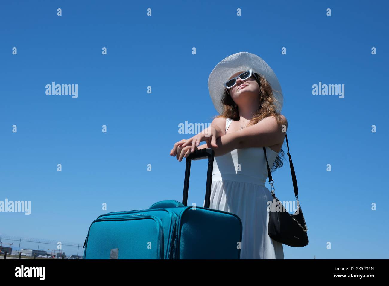 Le gambe di donna da vicino camminano su strada asfaltata e viaggiano con la valigia da viaggio . Autostop femminile in giro per il paese. Una giovane ragazza con un abito bianco e un cappello enorme con valigia viaggia in tutto il mondo Foto Stock