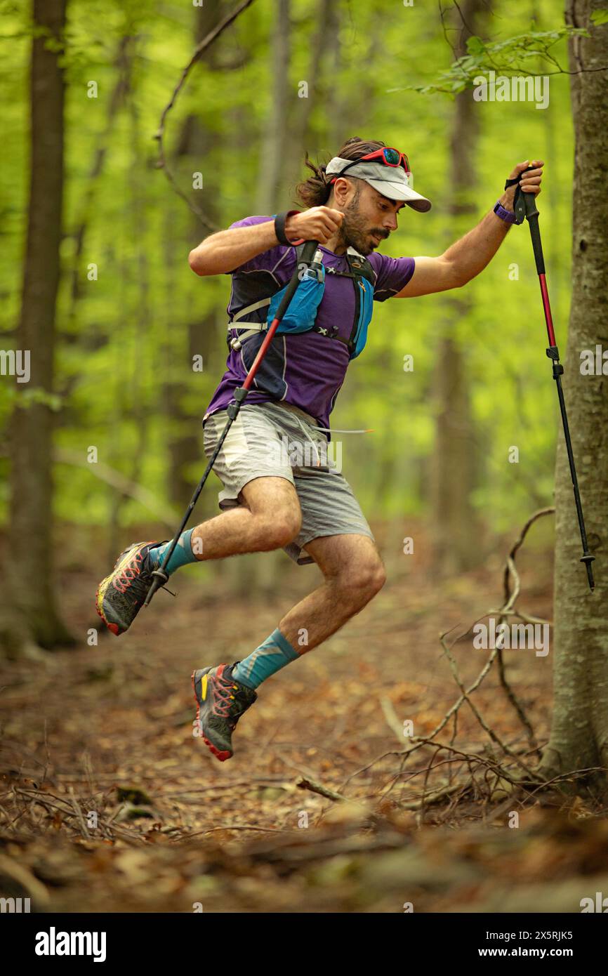 Un uomo con una camicia viola e pantaloncini grigi sta saltando sopra un albero tenendo due pali. Concetto di avventura e atletismo Foto Stock
