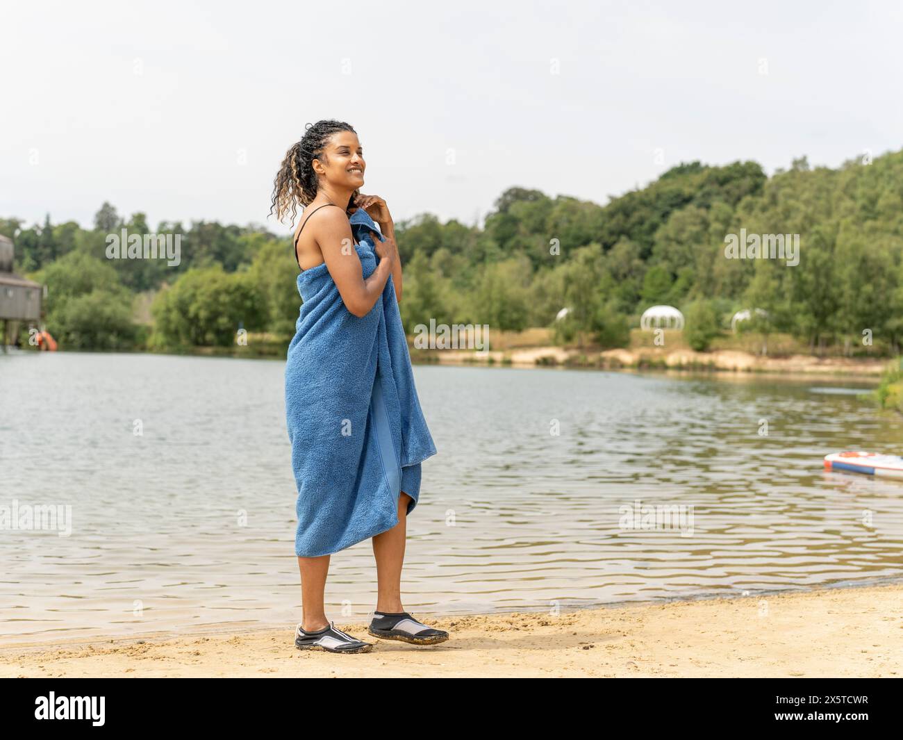 Donna sorridente in piedi avvolta in un asciugamano vicino al lago Foto Stock