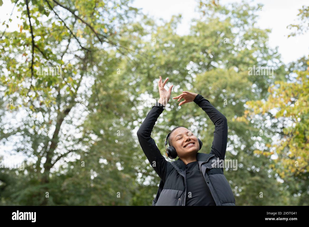 Ritratto di una donna sorridente con le cuffie che alzano le braccia nel parco Foto Stock