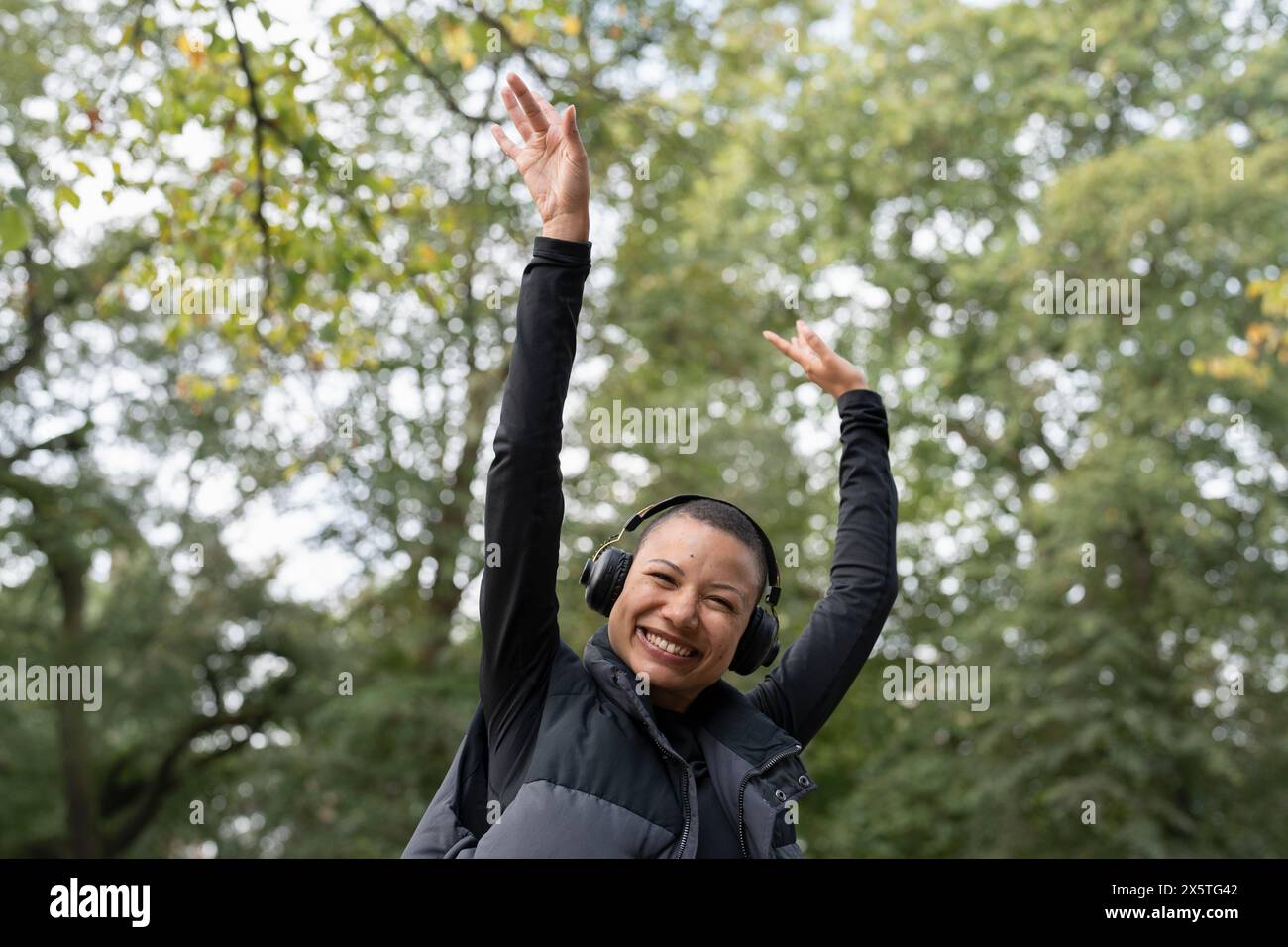 Ritratto di una donna sorridente con le cuffie che alzano le braccia nel parco Foto Stock