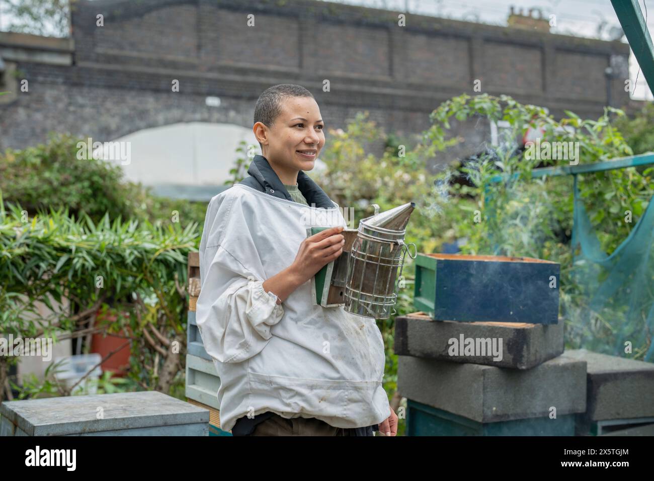 Donna sorridente apicoltrice che tiene il fumatore nel giardino urbano Foto Stock