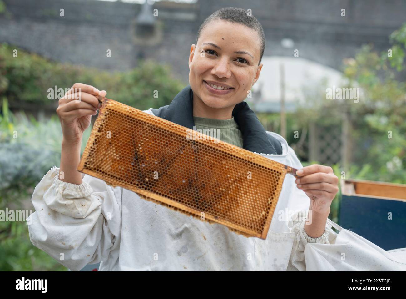 Ritratto di apicoltrice sorridente che tiene la cornice dall'alveare nel giardino urbano Foto Stock