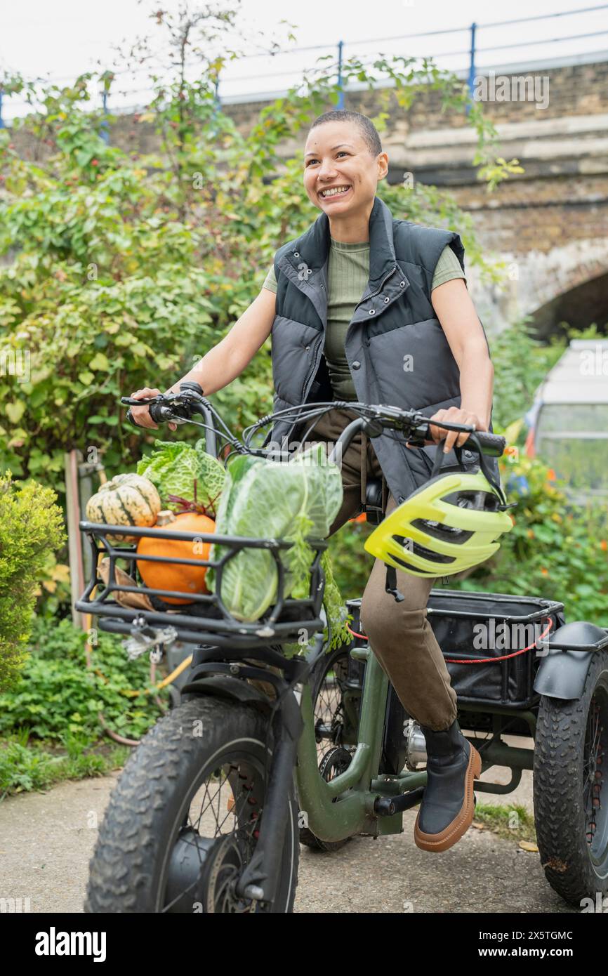 Donna sorridente che cavalca un trike elettrico carico di verdure fatte in casa Foto Stock