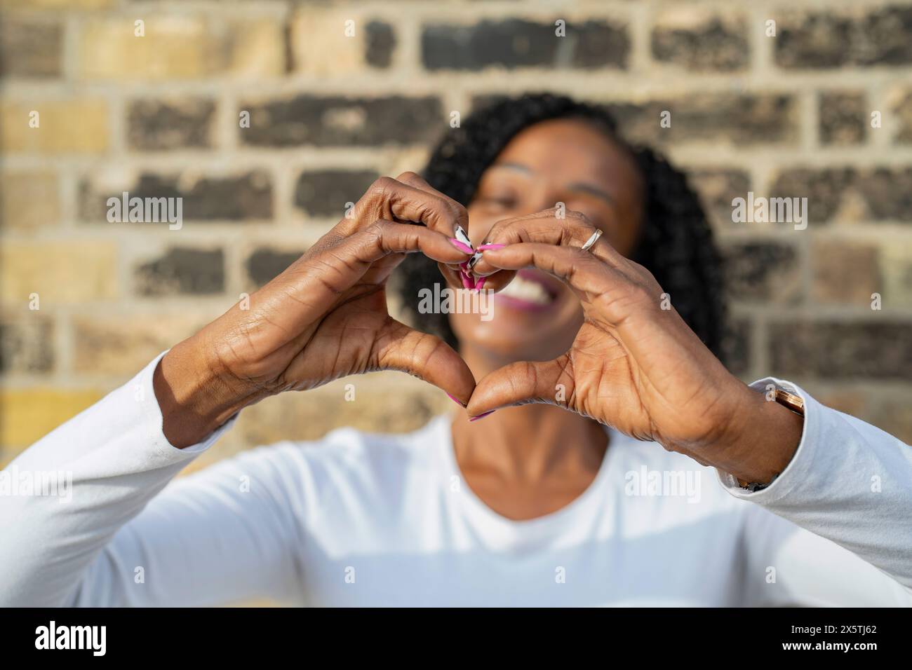 Donna che fa la forma del cuore con le mani contro il muro di mattoni Foto Stock