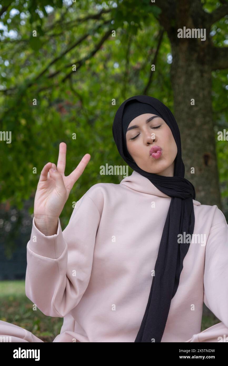 Ritratto di donna in hijab che fa volti e mostra un segno di pace nel parco Foto Stock