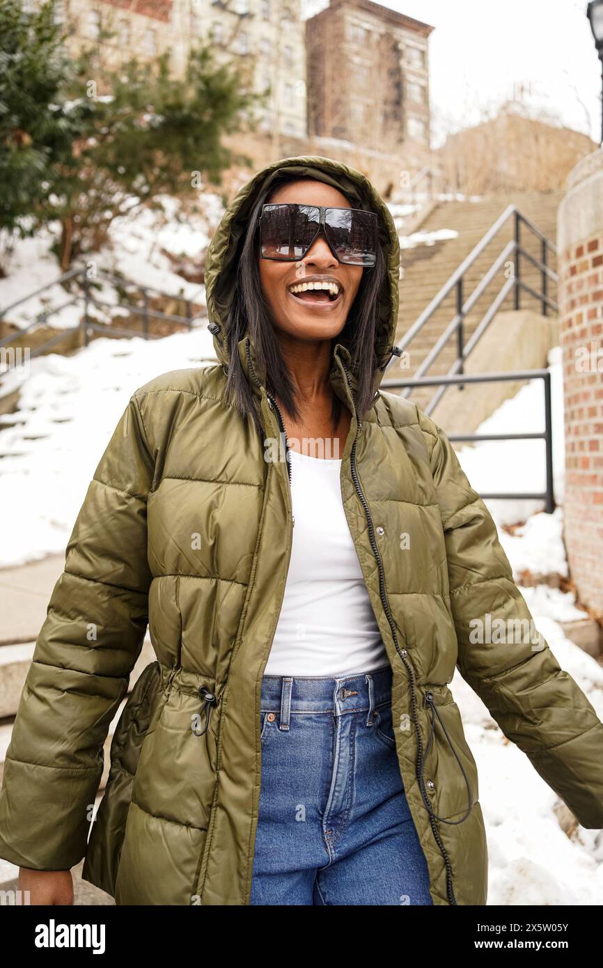 Donna sorridente in giacca invernale e occhiali da sole che cammina in città Foto Stock