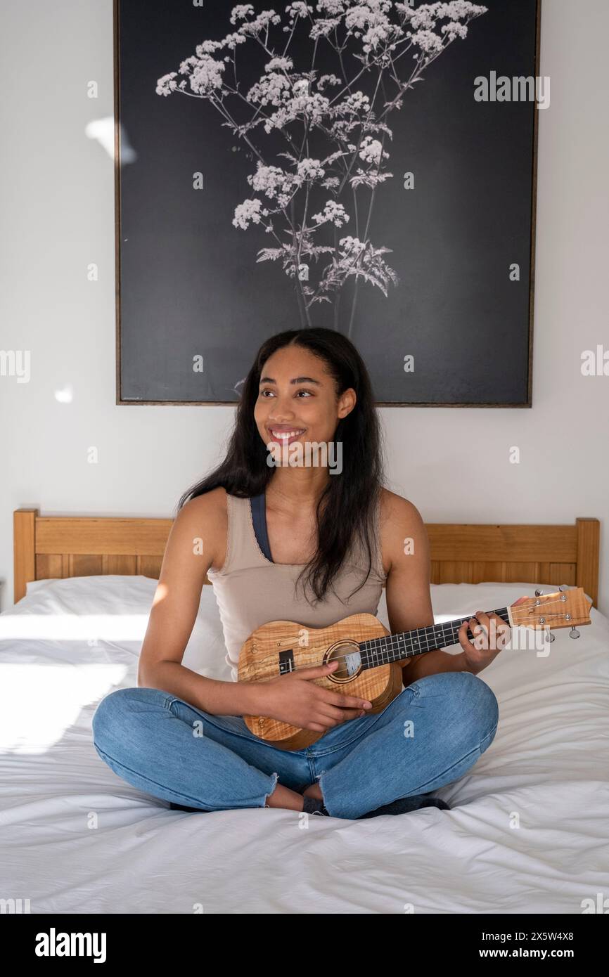 Giovane donna seduta sul letto a giocare a ukulele Foto Stock
