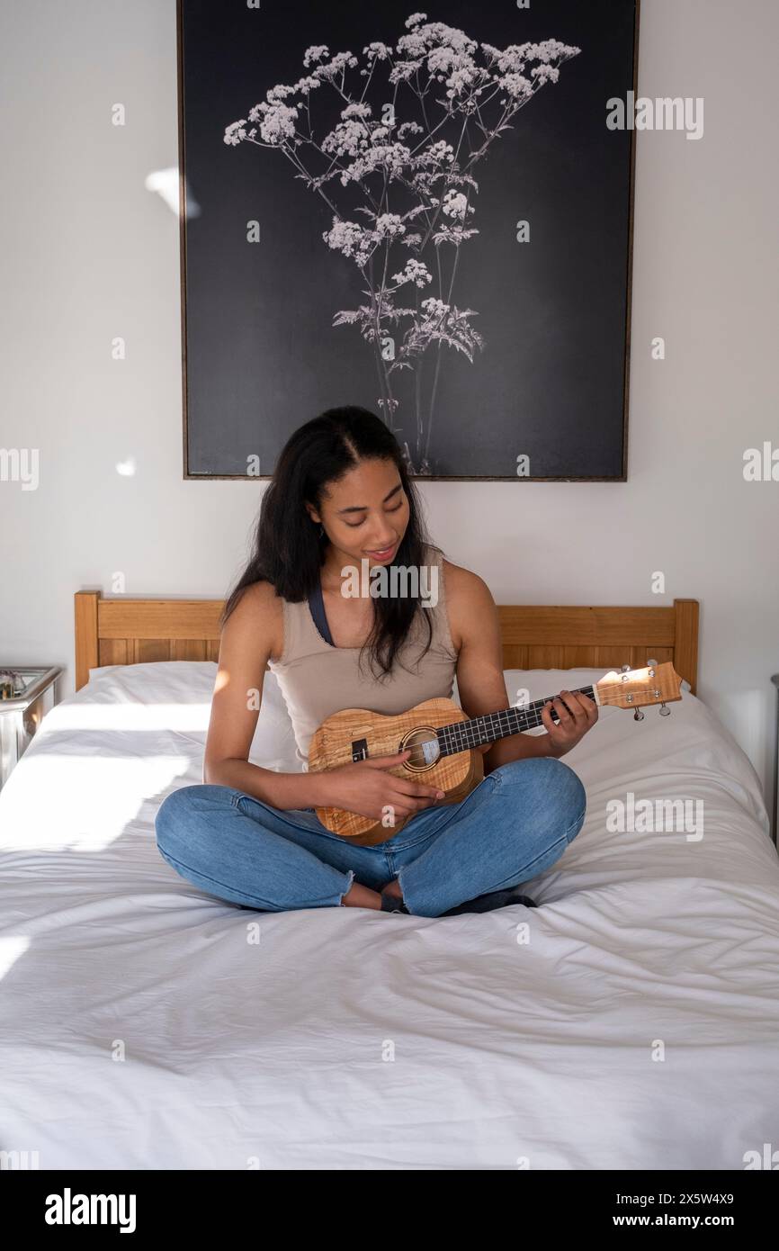 Giovane donna seduta sul letto a giocare a ukulele Foto Stock