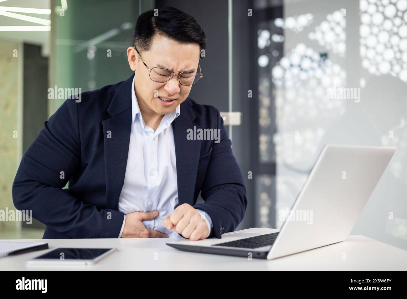 Un uomo d'affari asiatico di mezza età sente un improvviso dolore allo stomaco mentre si concentra sul lavoro su un notebook in un ambiente di lavoro elegante. Foto Stock