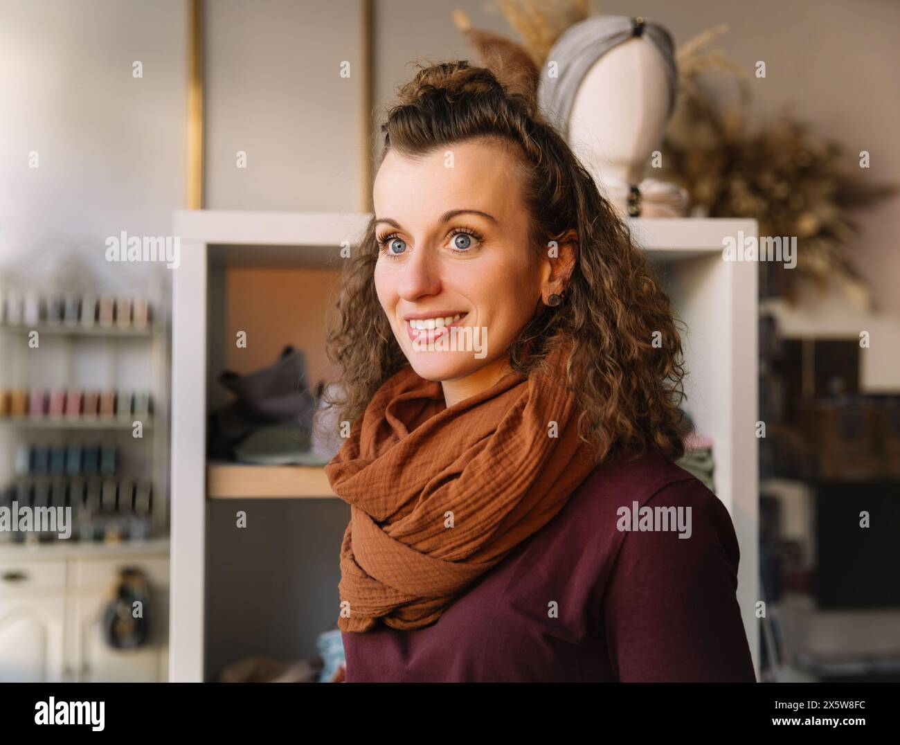 Donna dai capelli ricci che sorride dolcemente in un negozio di artigianato ben illuminato, circondata da materiali creativi e arredi eleganti Foto Stock