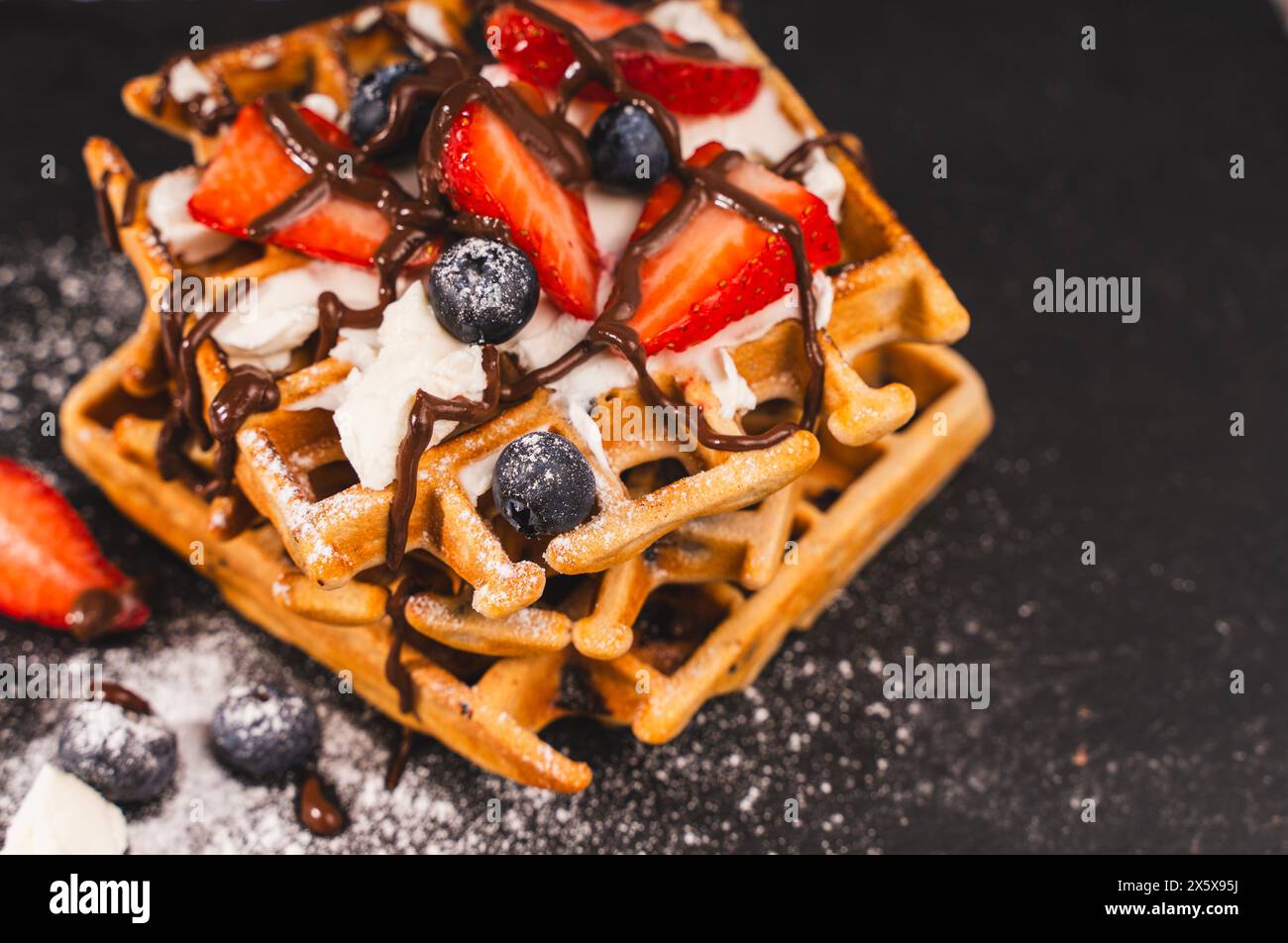 Primo piano del dessert. Waffle con crema di formaggio fresco, fragole e mirtilli, cosparse di zucchero a velo e condite con cioccolato su un baffo Foto Stock