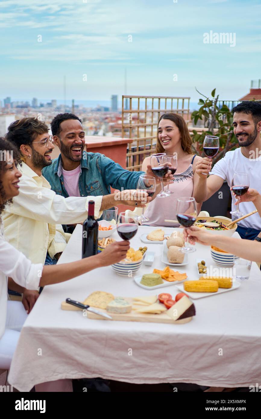 Gruppo di giovani felici che brindano vino rosso e festeggiano il pasto sul tetto Foto Stock
