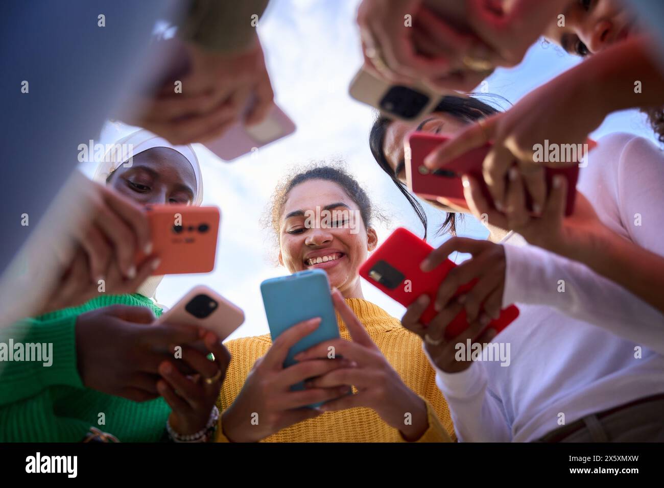 Angolo basso. Gruppo di giovani donne multirazziali sorridenti si riunirono in cerchio usando i telefoni cellulari Foto Stock
