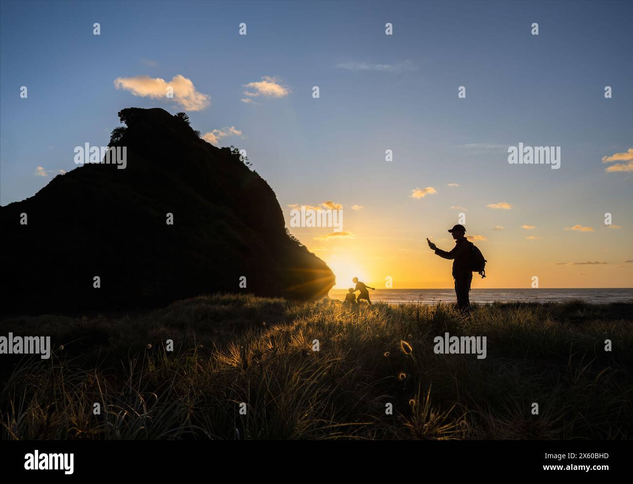 Uomo che scatta foto con uno smartphone di Lion Rock al tramonto. Bambini irriconoscibili che giocano a Piha Beach. Auckland. Foto Stock