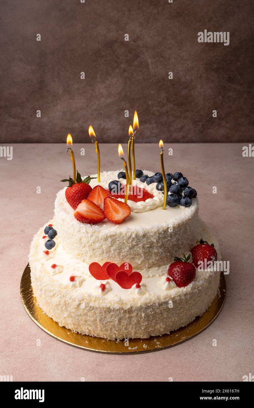 Torta di compleanno alla fragola e al cocco con panna e frutti di bosco freschi. Torta di compleanno a strati fatta in casa con candele accese. Messa a fuoco selettiva, primo piano. Foto Stock