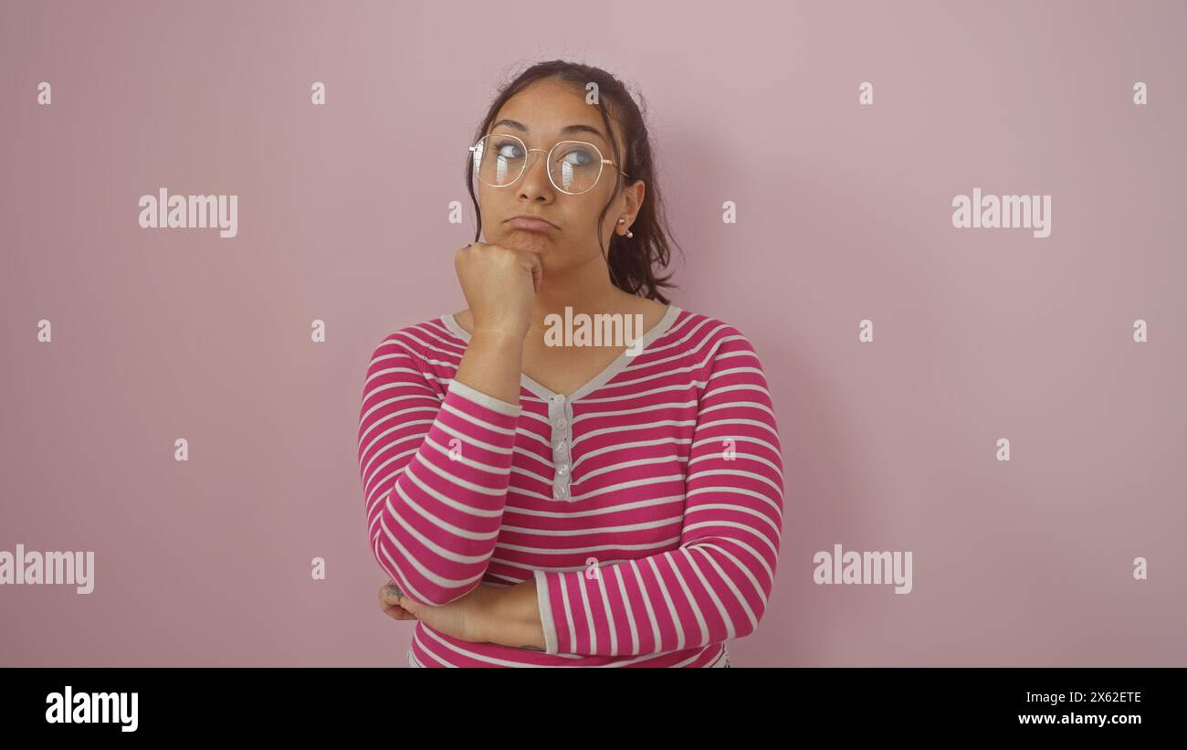 La giovane donna ispanica con gli occhiali, che indossa una camicia a righe contro una parete rosa, trasuda un atteggiamento premuroso. Foto Stock