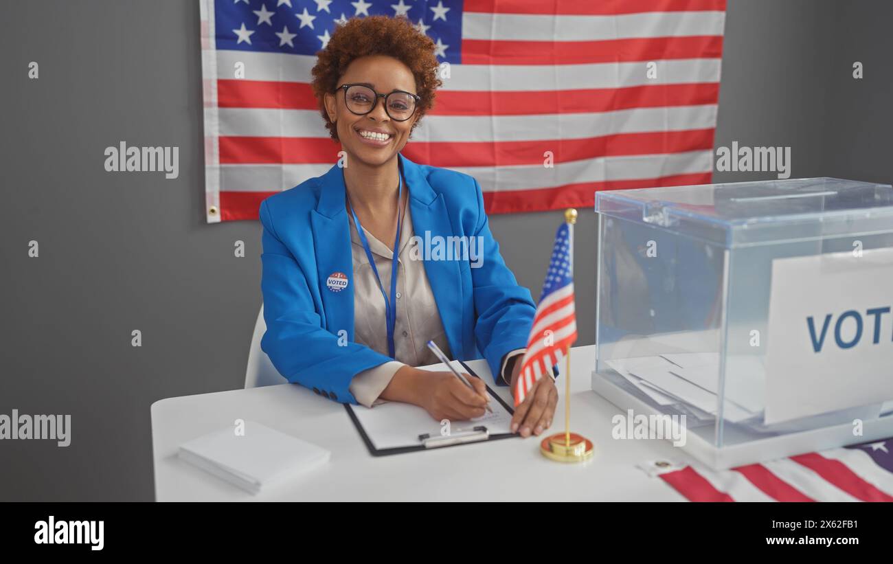 Una donna afroamericana sorridente in un blazer blu che prende appunti in un centro elettorale al coperto degli stati uniti con una bandiera americana. Foto Stock