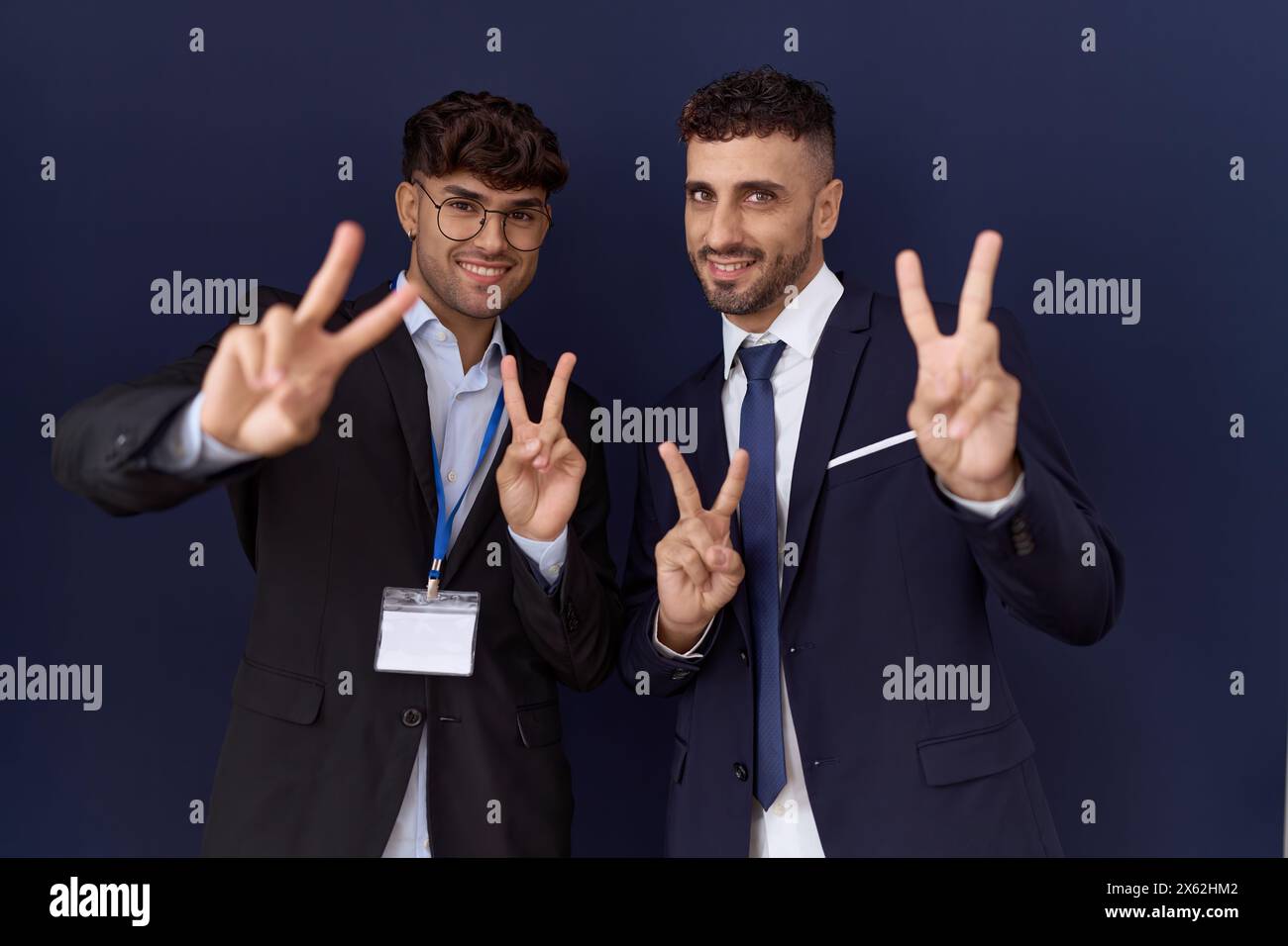 Due uomini d'affari ispanici che indossano abiti da lavoro sorridenti che guardano alla telecamera mostrando le dita che fanno il segno della vittoria. numero due. Foto Stock