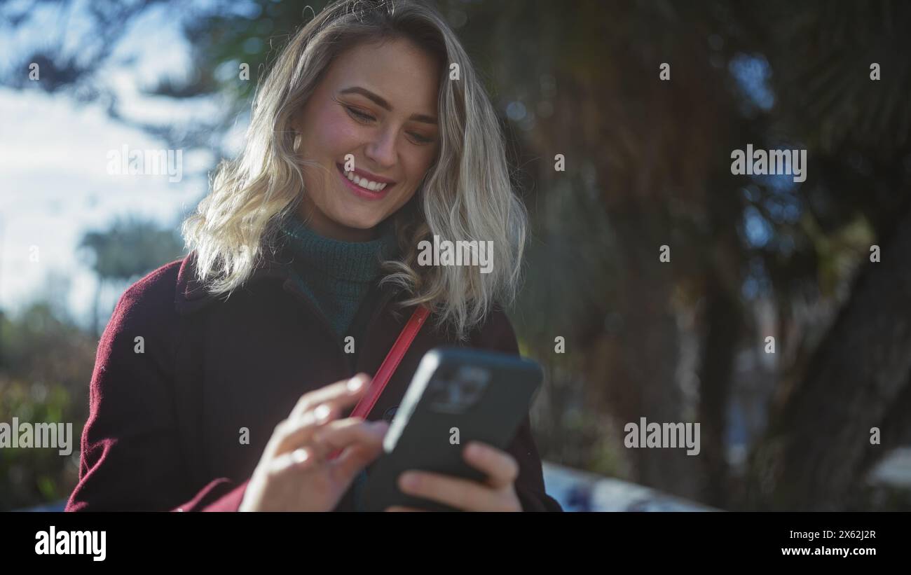 Una giovane donna sorridente con un cappotto Bordeaux che utilizza uno smartphone all'aperto in un parco soleggiato. Foto Stock