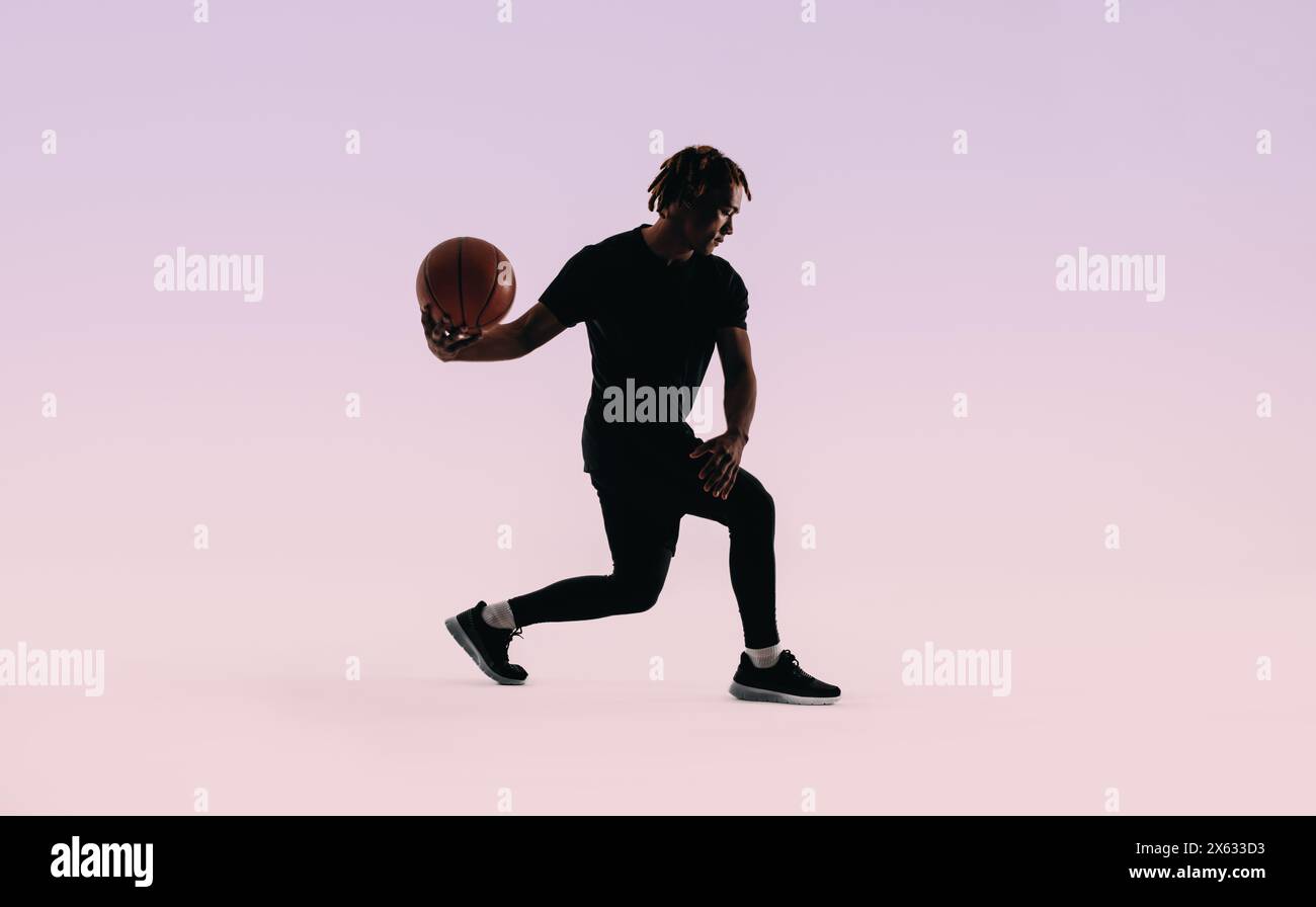 Sagomato sullo sfondo di un campo da basket, un giovane atleta con i dreadlock pratica il dribbling di una palla con una forma perfetta. Indossa un abbigliamento sportivo Foto Stock