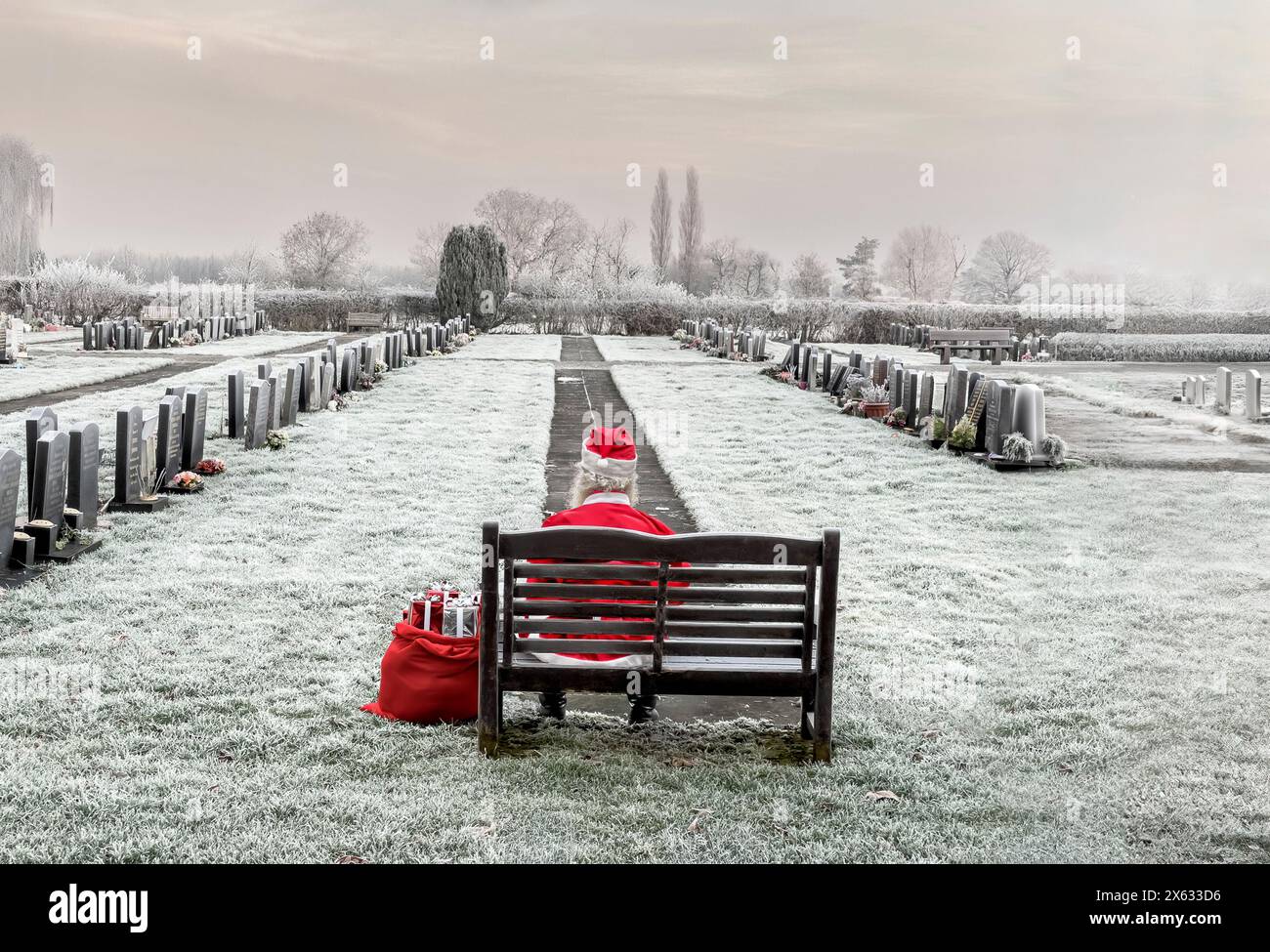 Una persona seduta da sola su una panchina di legno in un cimitero ricoperto di gelo, di nuovo alla macchina fotografica, vestita da Babbo Natale. REGNO UNITO Foto Stock