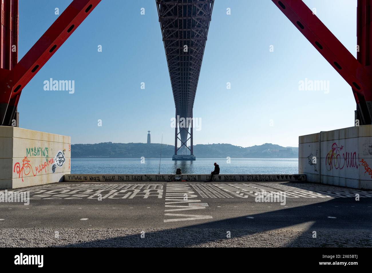 Sotto il torreggiante ponte 25 de abril, la struttura si estende lungo il fiume tago; una figura solitaria poggia sull'acqua su una sporgenza coperta di graffiti. Foto Stock