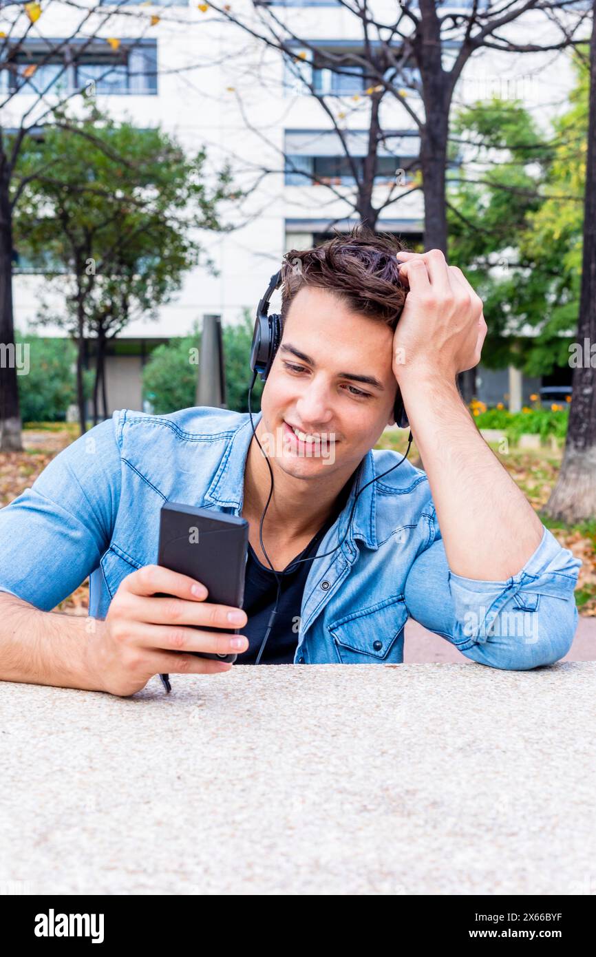 Un giovane allegro in denim si appoggia alla mano mentre utilizza lo smartphone e le cuffie, seduto in un parco urbano con alberi e un edificio moderno sul retro Foto Stock