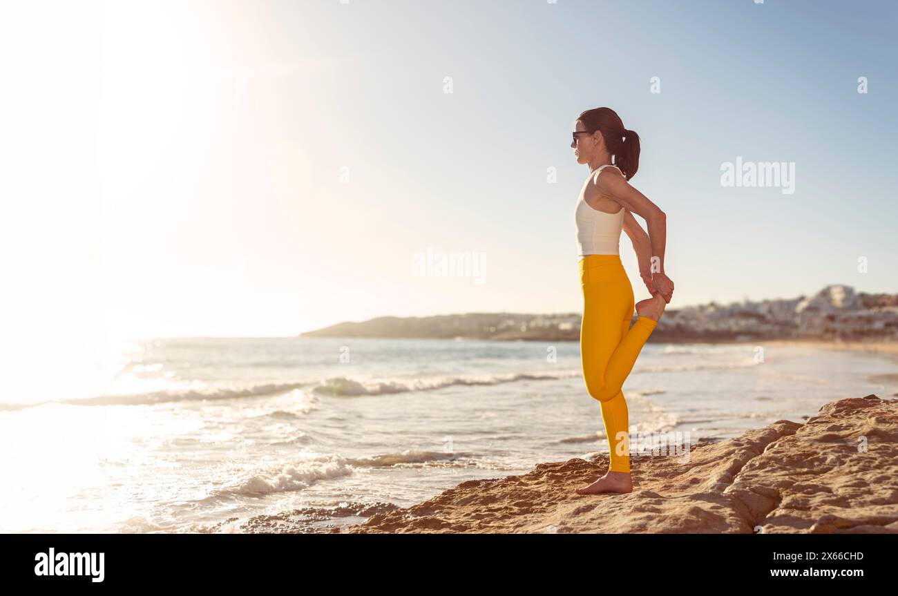 Donna sportiva che fa un esercizio di riscaldamento delle gambe in riva al mare Foto Stock