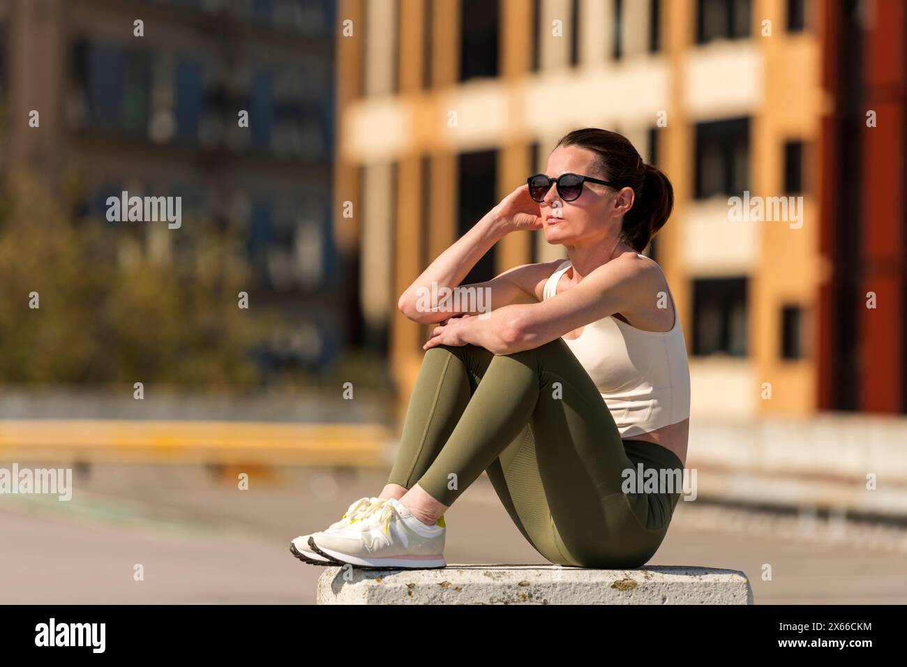 Donna sportiva seduta riposante al sole dopo l'esercizio fisico all'aperto, sullo sfondo urbano Foto Stock