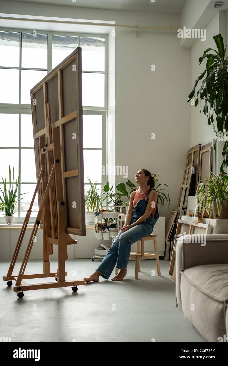 Donna allegra artista che guarda enormi tele ammirando mentre si esamina seduto in uno studio d'arte domestico. Foto Stock